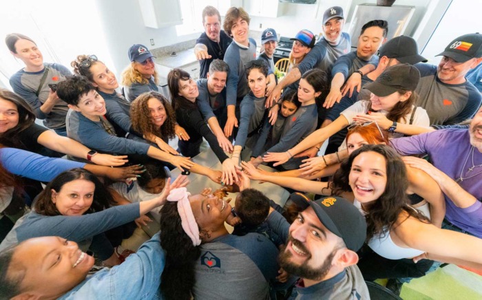 A diverse group of over 20 volunteers gathers in a bright, modern kitchen for a celebratory huddle. Taken from a high-angle perspective, the image shows the team reaching toward the center in a unified 