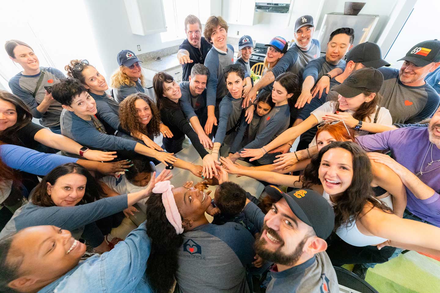 A diverse group of over 20 volunteers gathers in a bright, modern kitchen for a celebratory huddle. Taken from a high-angle perspective, the image shows the team reaching toward the center in a unified "hands-in" gesture. Many participants wear matching blue and grey apparel featuring a heart logo, their faces filled with smiles and shared excitement. This vibrant scene captures the spirit of community, collaboration, and collective impact.